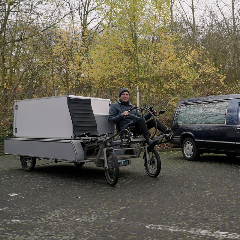 Jürgen Dahlfeld auf dem Bestattungsfahrrad. Daneben ein Leichenwagen.
